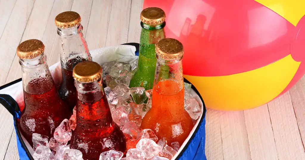 Closeup of a cooler with soda bottles and ice on a rustic wood table with a beachball in the background.

