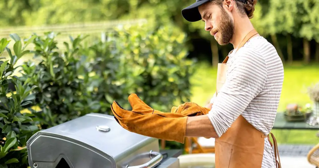 Handsome man in leather apron and gloves preparing for cooking on the modern gas grill at backyard of country house