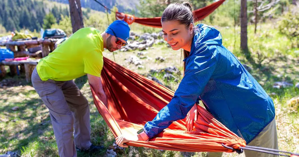 Man and woman in hiking equipment setting up a hammock between trees at the top of the mountain.