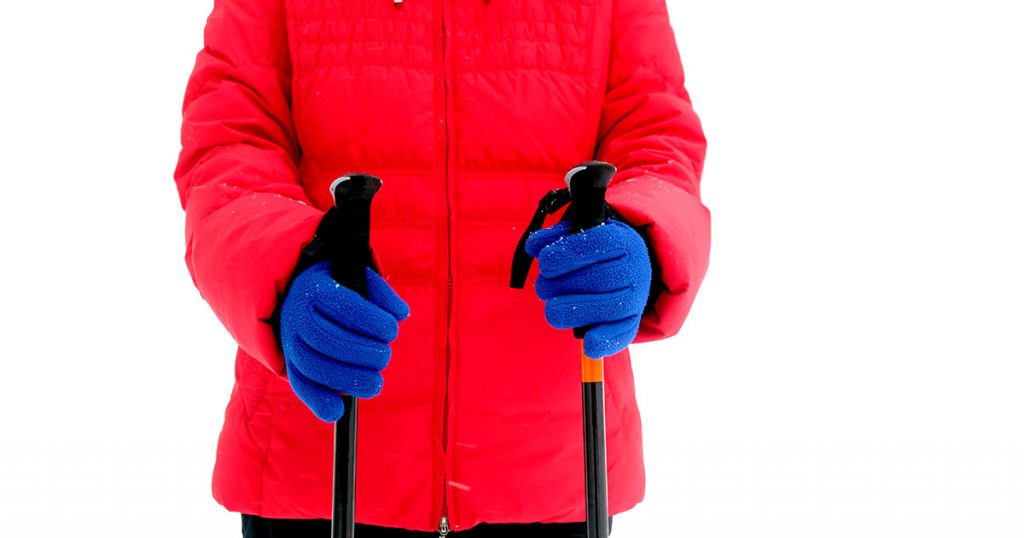 Woman in red sport jacket and blue gloves holds two sticks for hiking in winter