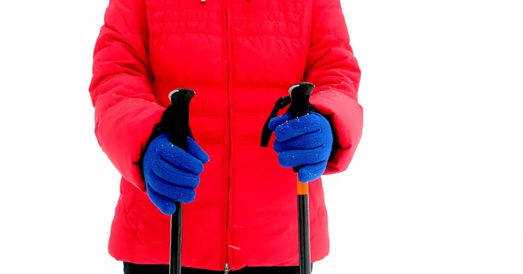 Woman in red sport jacket and blue gloves holds two sticks for hiking in winter