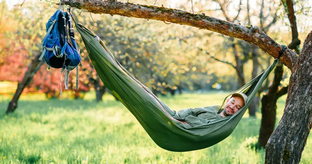 man in green hat lying in hammock on apple tree at blooming garden in summer sunny day on vacation