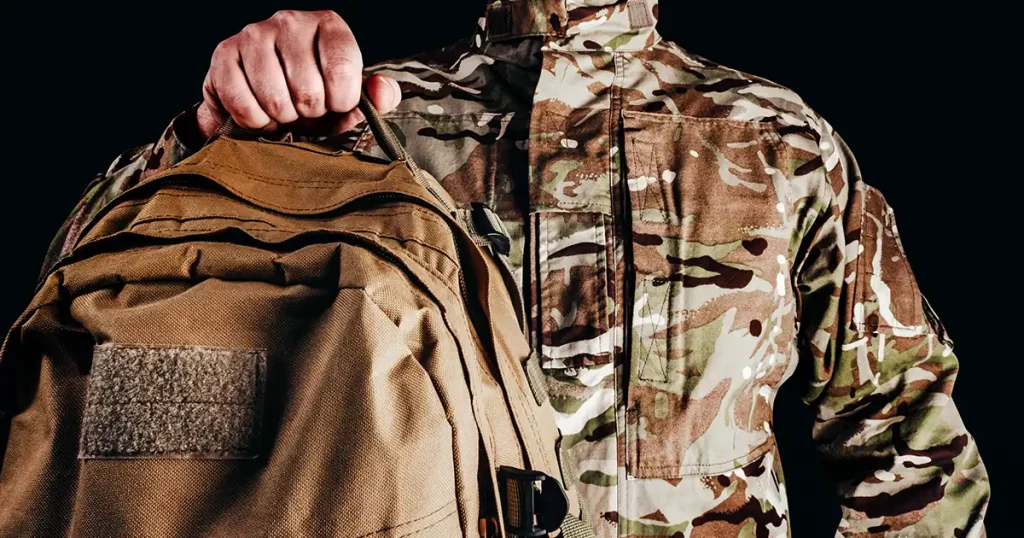 Photo of soldier in camouflaged uniform and tactical gloves holding olive colored backpack on black background.