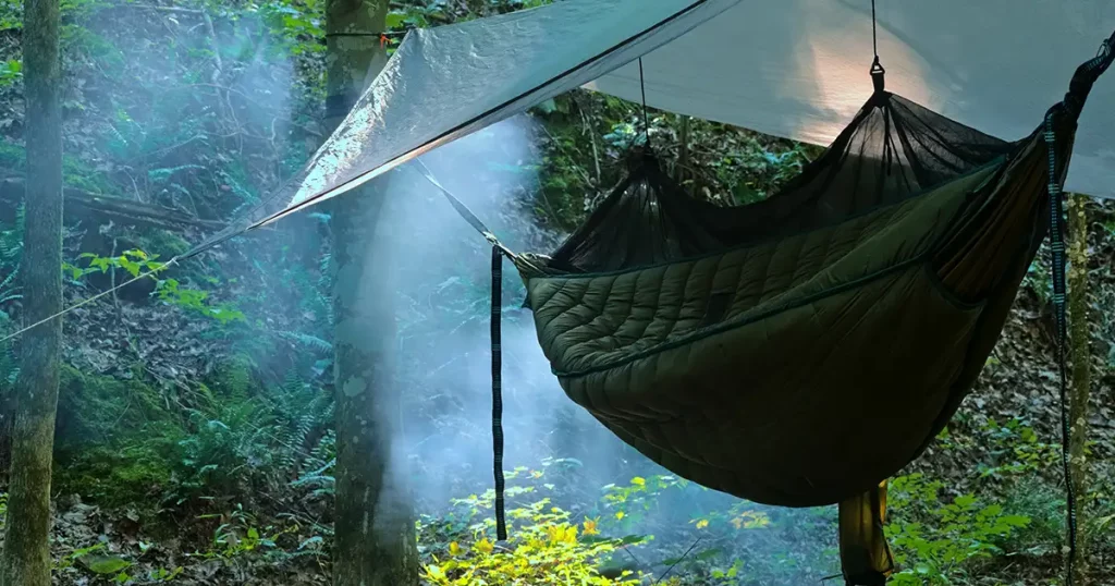 Hammock with a bug net set up between two trees underneath a tarp with campfire smoke bellowing behind it.