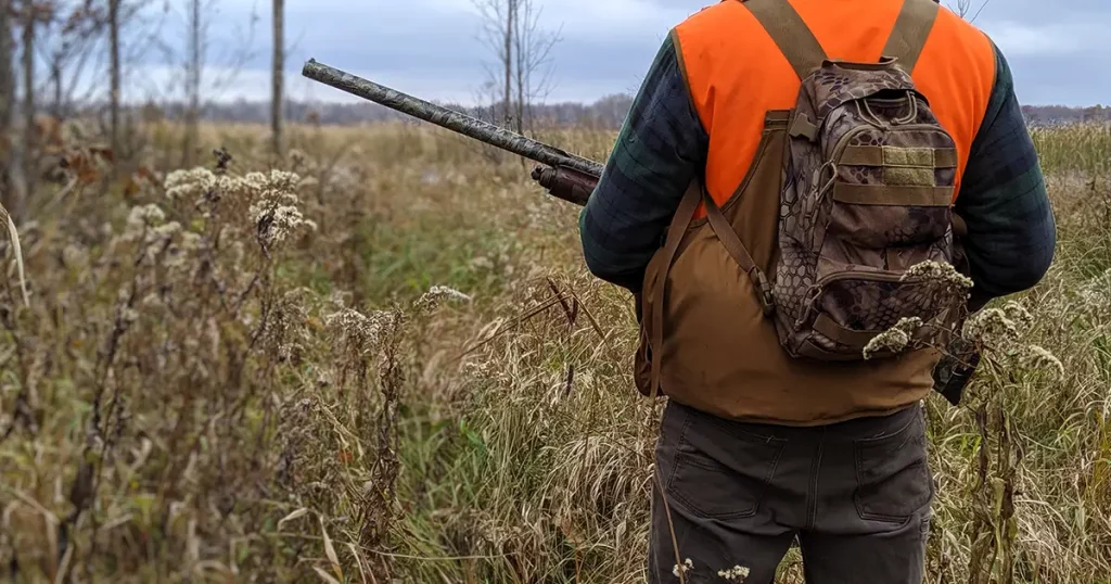 Man holding shotgun wearing bird hunting vest and camouflage backpack in outdoor scene