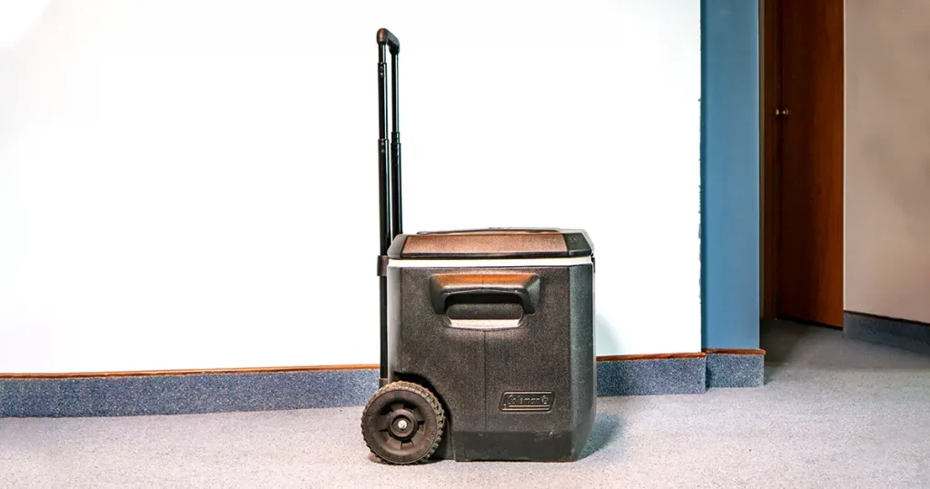 A black Coleman cooler sits on the floor in an apartment room, packed for a trip