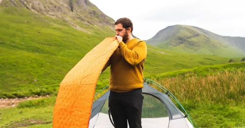 Young and attractive man inflating a mattress next to his tent to do solo camping in Scotland
