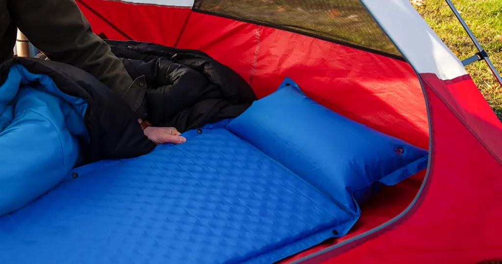 A man gets his tent and sleeping bag ready at a campground by inflating and setting up his blue blow-up mattress pad to put for under his sleeping bag