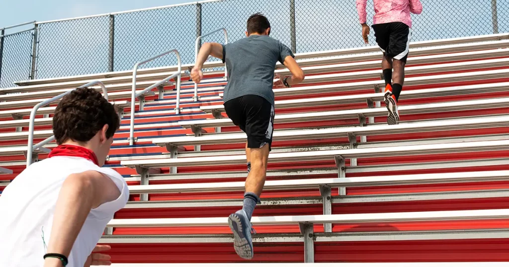 Rear view of three high school boys running up red stadium bleachers durint track practice.
