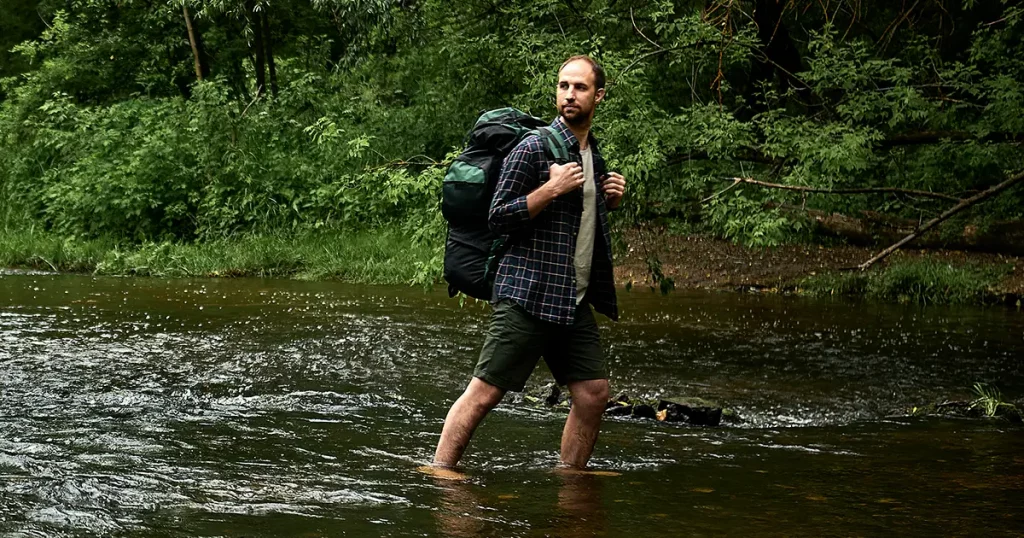 A man in a plaid shirt and green shorts with a large hiking backpack looking back at the far bank of the rive