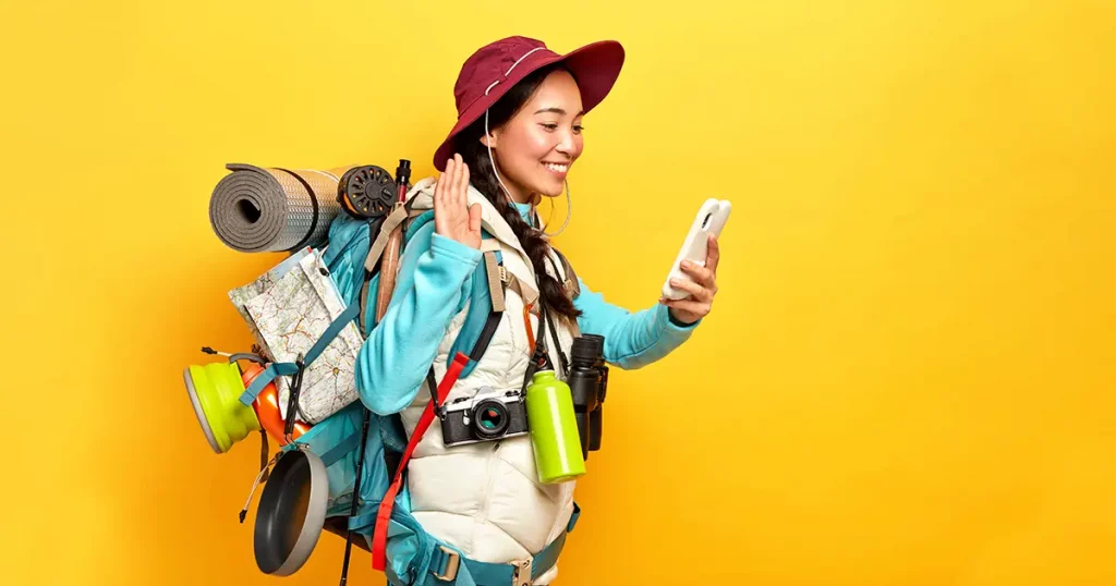 girl waves palm, greets someone via video call, holds mobile phone in hand, carries rucksack with all necessary thing