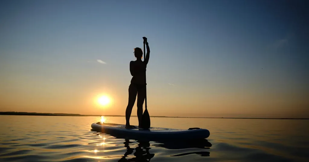 Siluet of Woman standing firmly on inflatable SUP board and paddling through shining water surface.