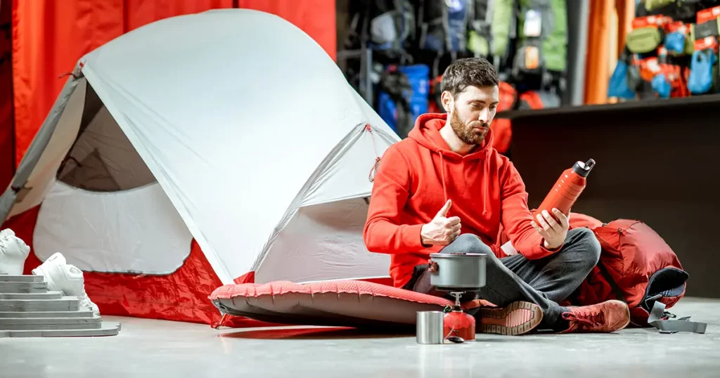 Man trying camping tools sitting on the floor with travel equipment and tent on the background in the shop