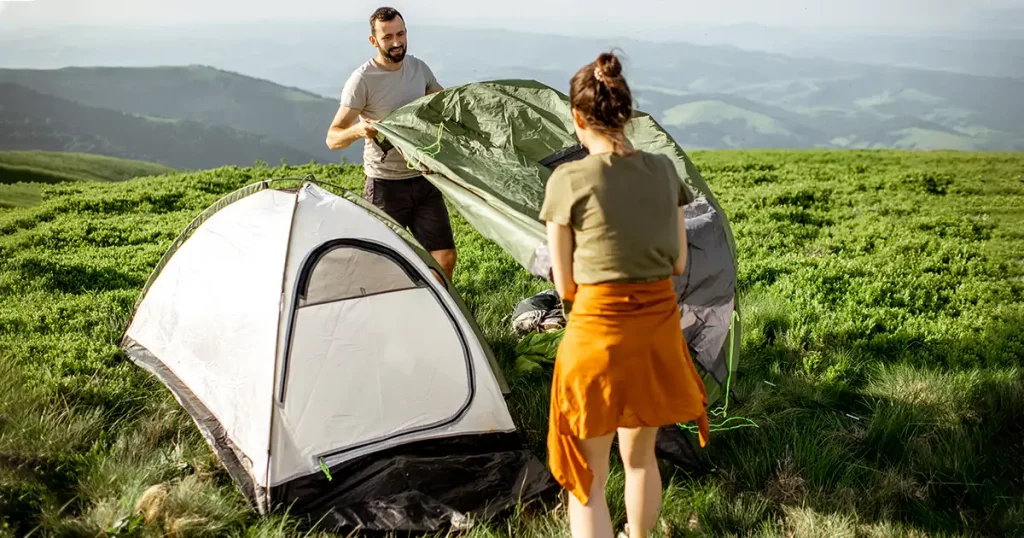 Young couple setting up the tent on the green meadow, traveling high in the mountains during the sunset