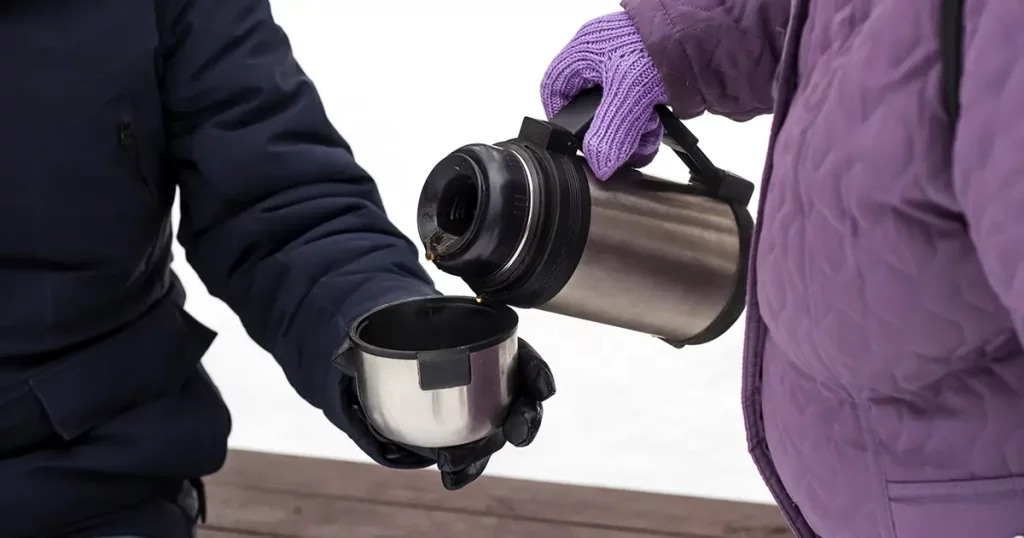 a woman pours tea from a thermos into a thermos cup for a man to drink and keep warm.