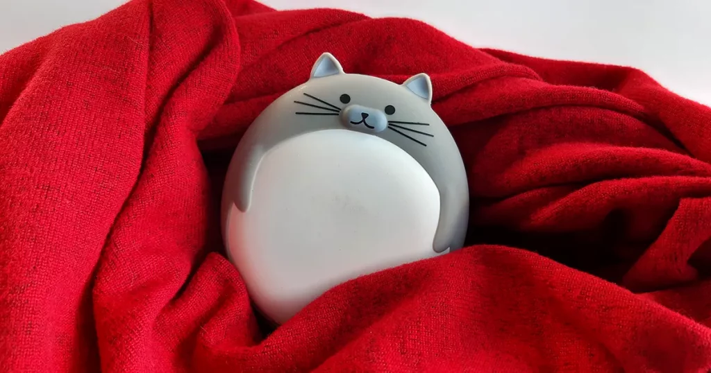Hand warmer in the form of a round gray-white cat on a red woolen scarf on a white background.