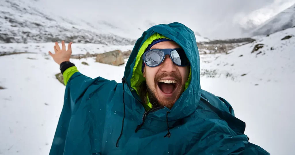 Selfie of smiling hiker in sunglasses and blue raincoat poncho in snowy mountains
