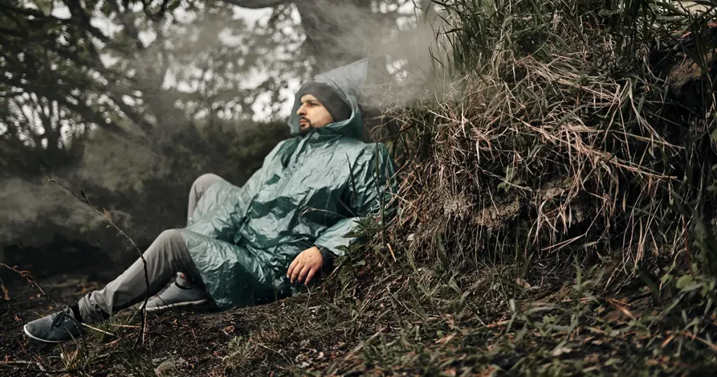 A man with a beard in a green raincoat sits on the ground in nature