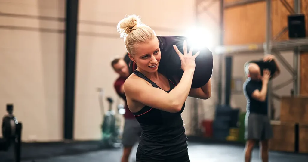 Fit young woman in sportswear working out with a weight bag over her shoulder during an exercise class at the gym