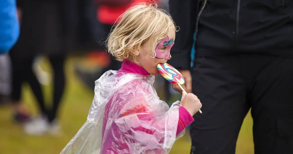 little girl with make up on her face having a rainbow lollipop on the festival field Traenafestival