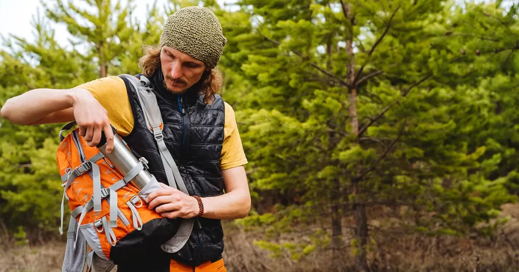 Metal bottle with water, a person puts a thermos in the pocket of a backpack