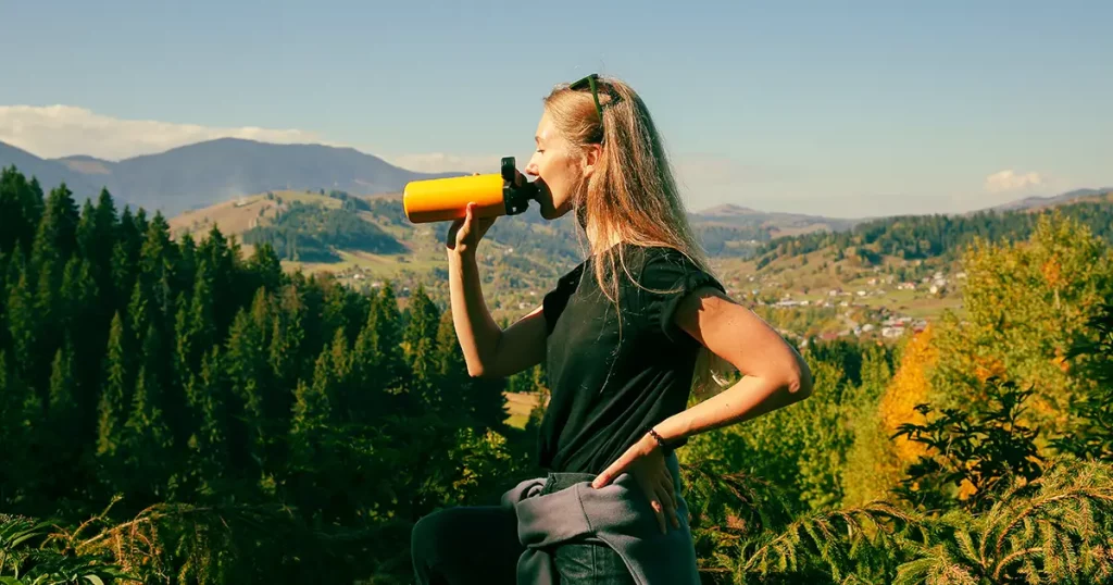 woman with long straight blonde hair, wearing black t-shirt, drinking from yellow thermo bottle, gorgeous landscape of mountains covered with coniferous forest